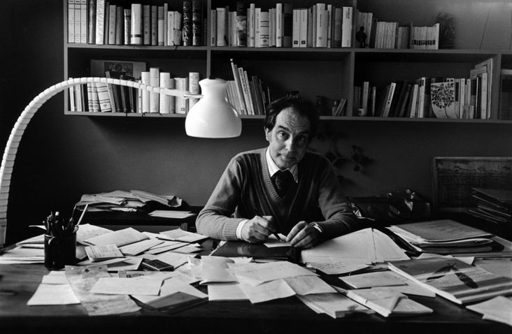 fotografia mostra Italo Calvino sentado em escrivaninha repleta de papeis, com estantes de livros na parede atrás